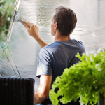 A man cleaning a glass barrier outdoors with a squeegee, surrounded by greenery.