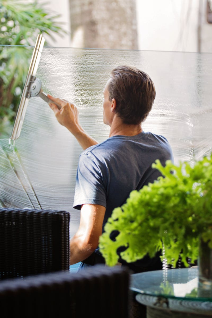 A man cleaning a glass barrier outdoors with a squeegee, surrounded by greenery.